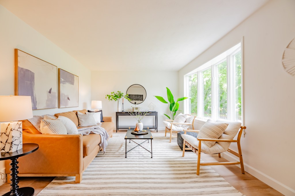 Bright and airy living room featuring a brown sofa, two armchairs, and a coffee table, with large windows allowing natural light and green plants adding freshness. Updated Cape Cod Home in Penfield