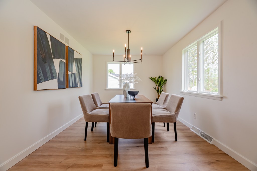Modern dining room featuring a wooden table, beige upholstered chairs, and abstract wall art, with natural light from windows. Updated Cape Cod Home in Penfield